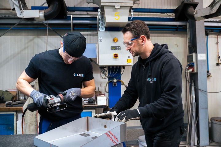 Zwei Personen bearbeiten ein Metallteil in einer Werkstatt mit einem Schleifgerät.